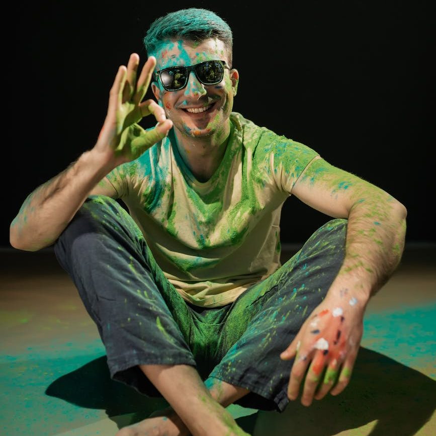 a man with holi powder wearing sunglasses sitting on floor giving an ok sign